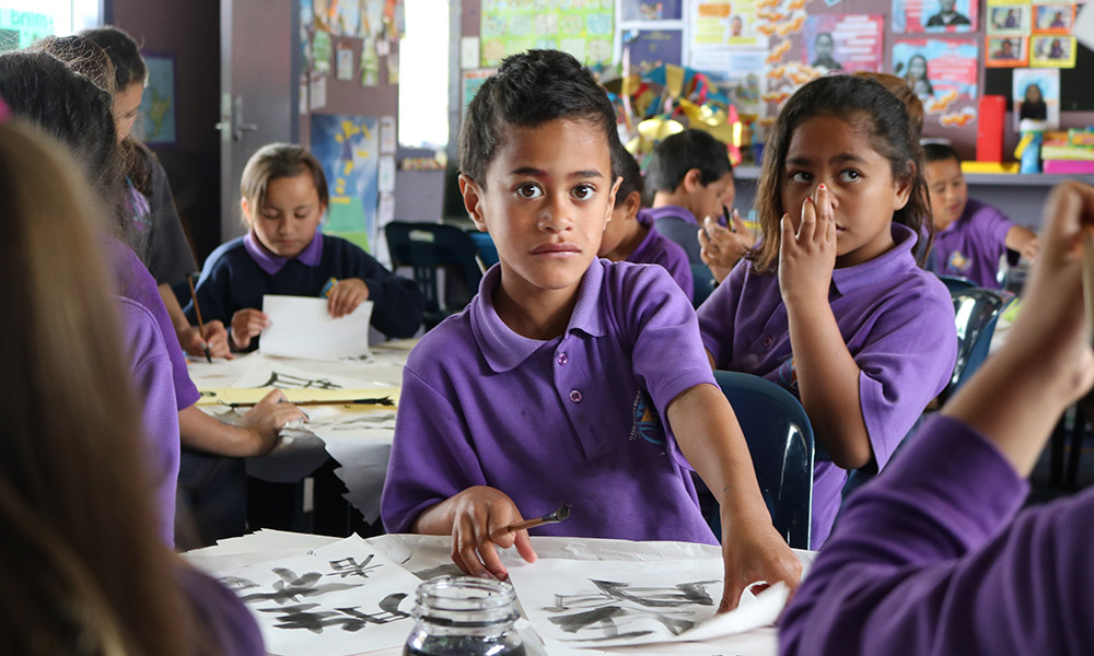 A boy surrounded by other kids sitting at tables practicing calligraphy in a school room