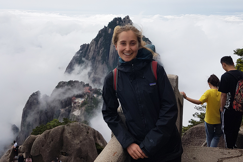 Cleo leaning on a handrail with a rocky mountaintop peaking through the clouds behind her