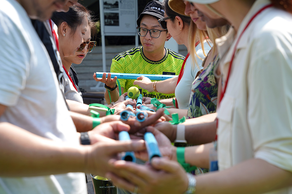 People playing a game where they have to transfer a tennis ball between each other by rolling it along plastic pipes and eventually getting it to a bucket