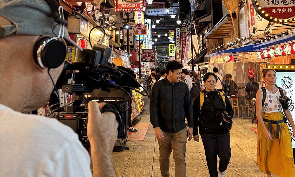 A cameraman filming three people walking through a busy walking street in Japan at night, lit up by shop signs and lanterns