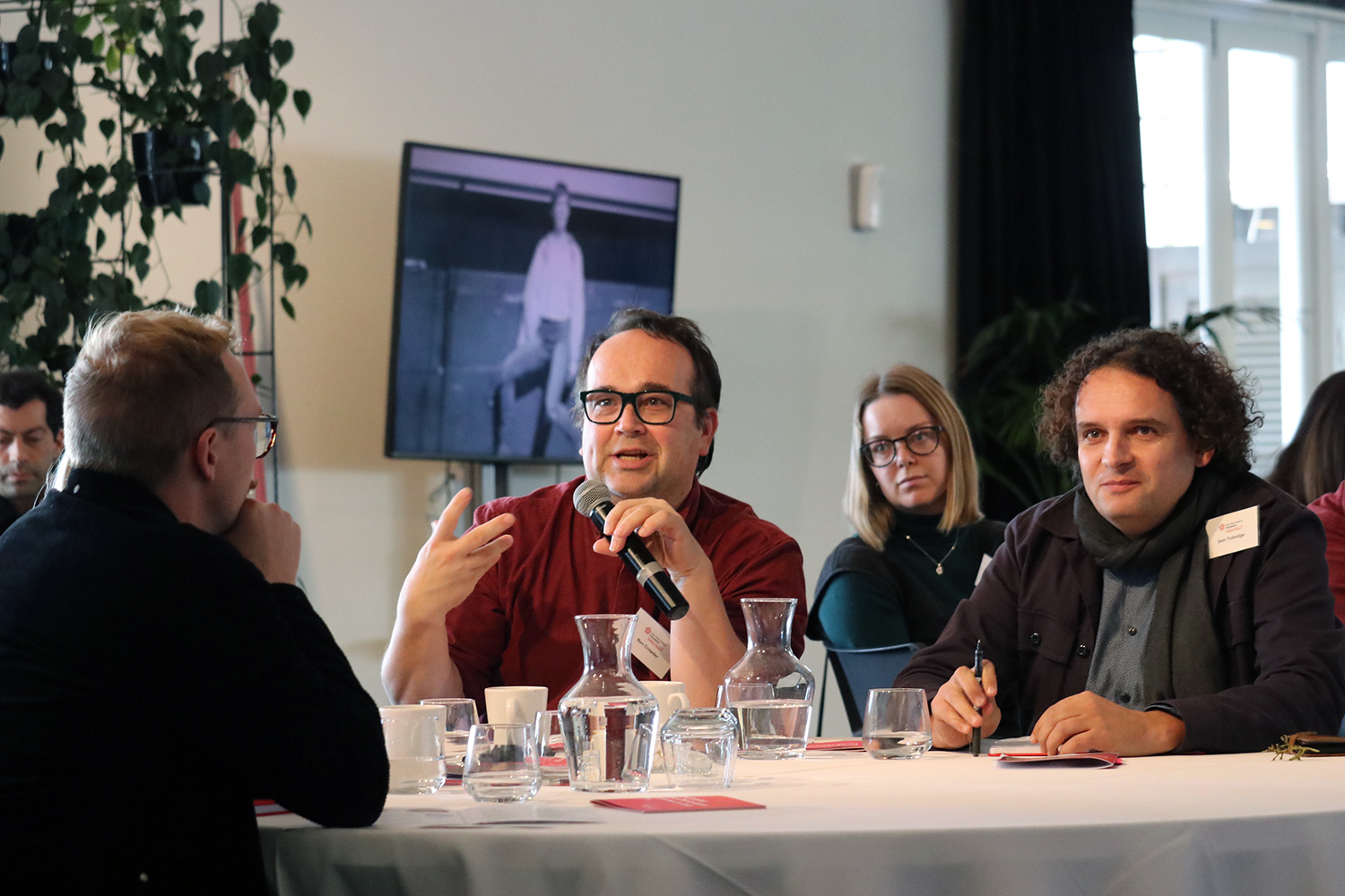A man speaking into a microphone while sitting at a table with two others