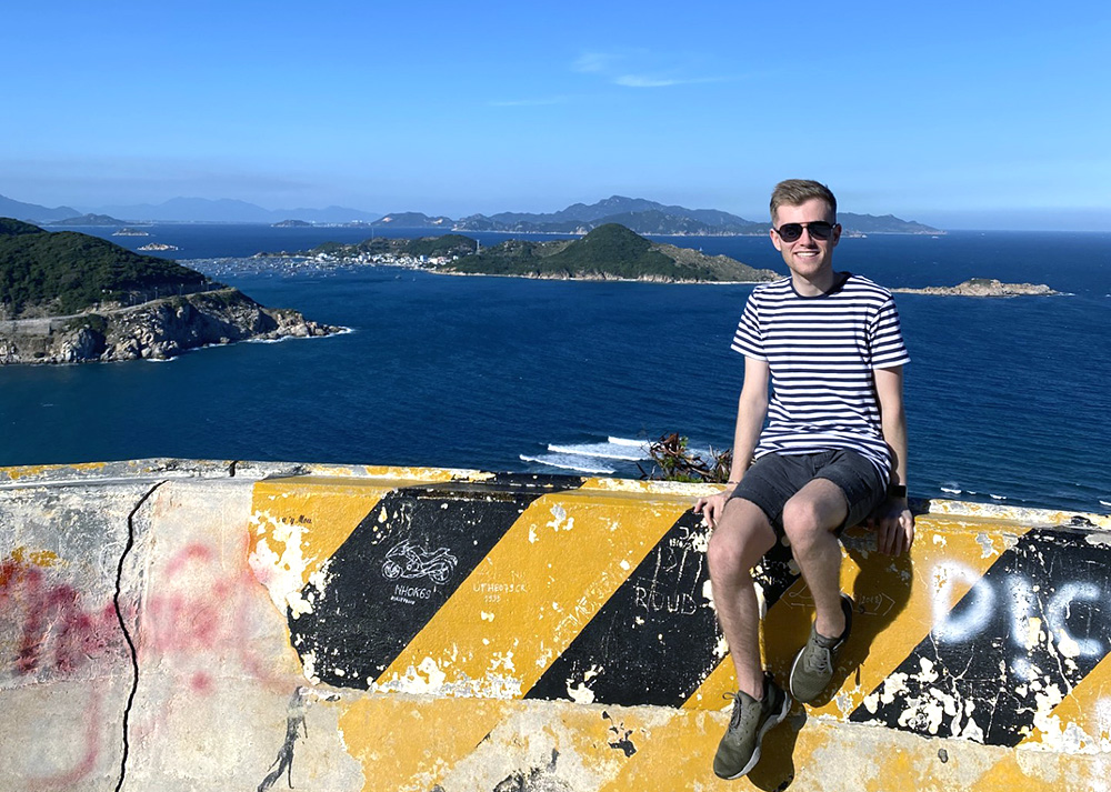 Ethan sitting on a bollard with a view of the sea and coast behind him