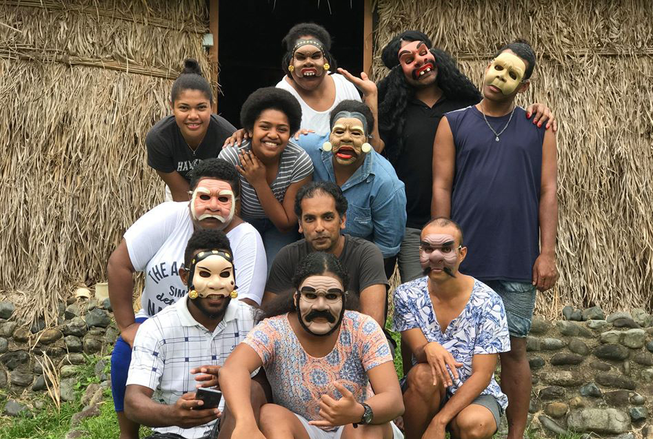 Jacob crouching in front of a thatched building with a group of people wearing masks