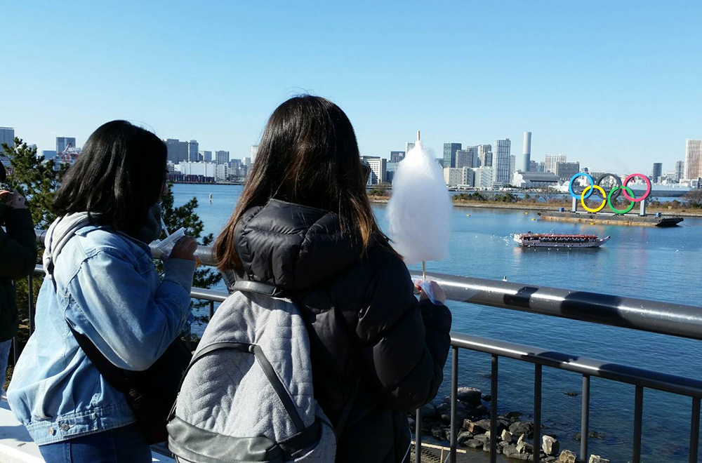 Varenica Gunawan and a friend looking out over a body of water with a ferry on it and the Olympic rings visible on an island