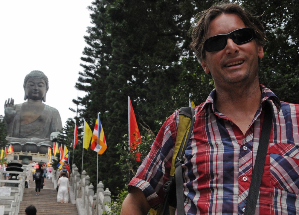 Man smiles a bottom of stairs, leading up to large buddha statue