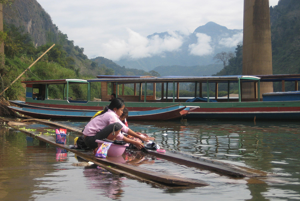 A woman washing clothes in a river with wooden boats behind her