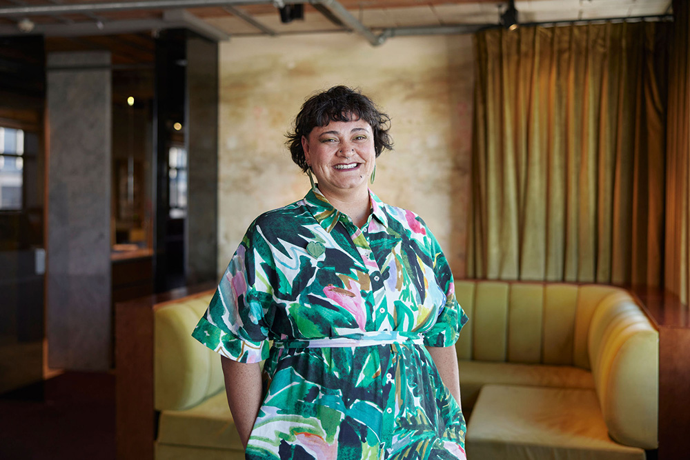 Sarah in a floral green jumpsuit standing in a mustard-coloured room