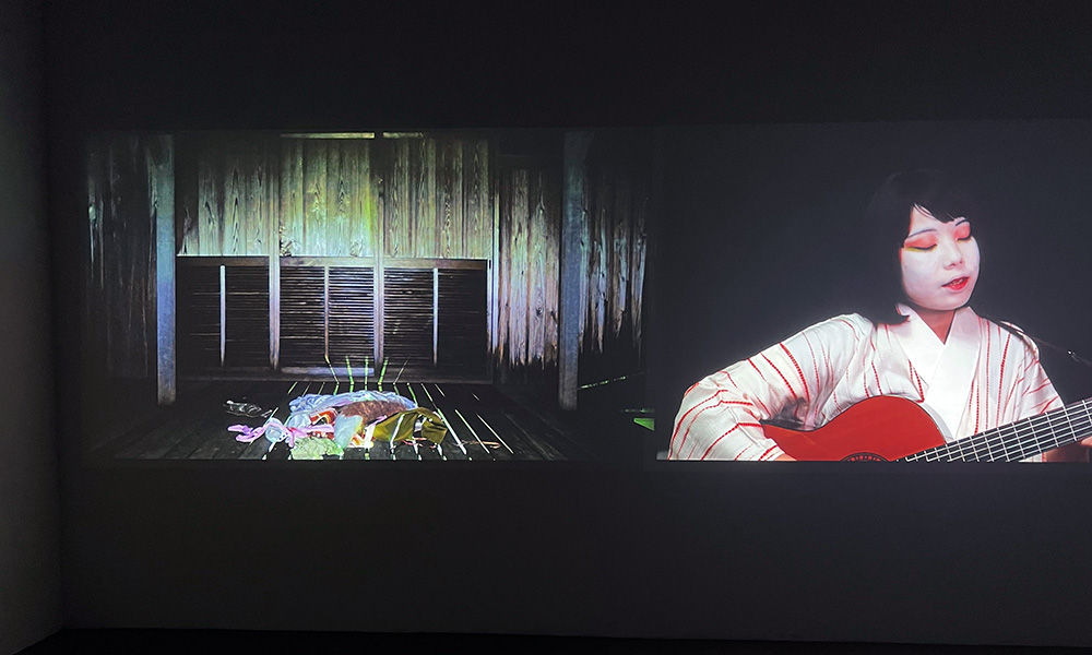 A photo of a work of art depicting a woman in traditional Japanese clothing and face paint playing a guitar and a room with a pile of clothing in the middle