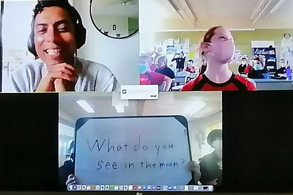 A screenshot of a Zoom call with three participants - a man, a girl and students holding a sign saying what do you see in the moon