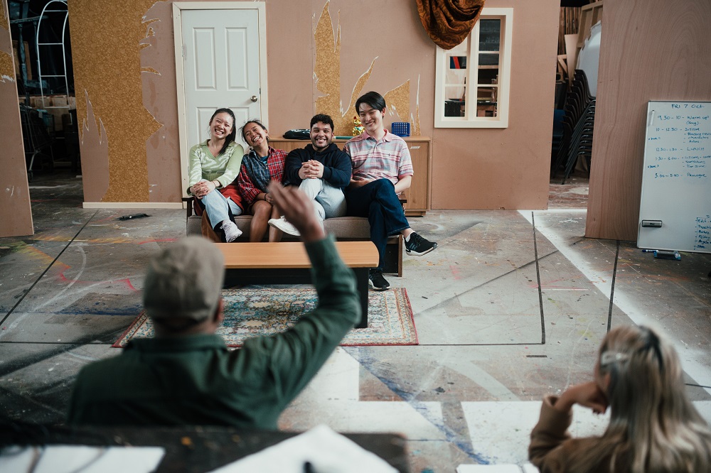 A theatre set made to look like a sitcom set with four people sitting on a couch watched on by a man waving his hand in the air