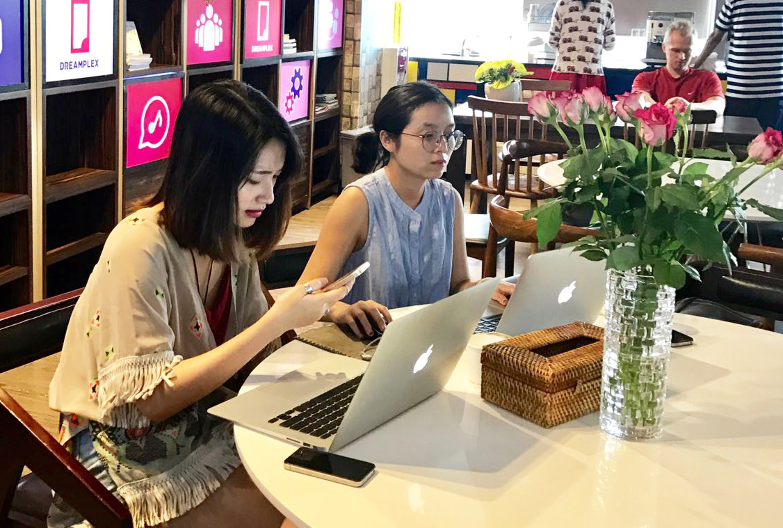 A woman looking at her cell phone as a woman beside her works on a laptop