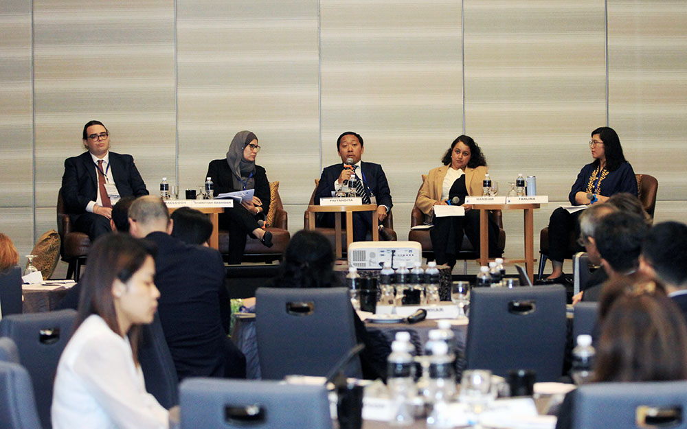 A fiv-person panel sitting in front of a seated audience, one of them speaking into a microphone