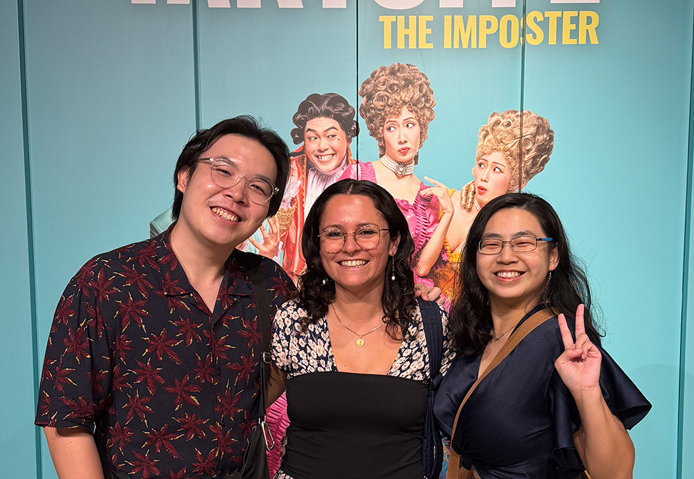 Monica and two colleagues posing for a photo in front of a poster for the film The Imposter
