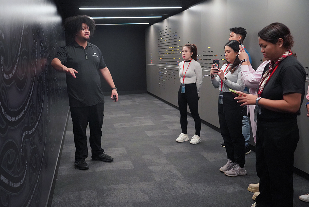 A guide in an All Blacks jersey speaking to the group in front of a large artwork that takes up a wall