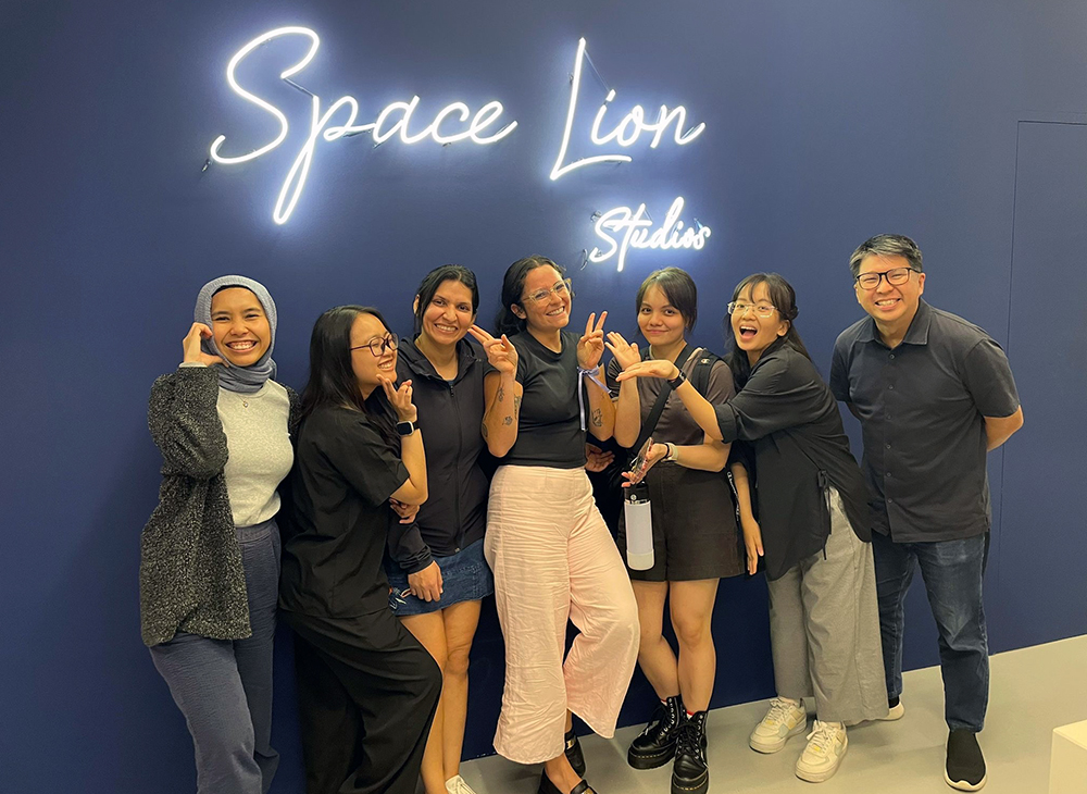 Monica posing for a photo with colleagues in front of a neon sign reading 'Space Lion Studios'