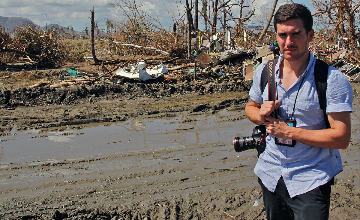 A journalist in the field on a mud road with storm damaged trees and structures behind him