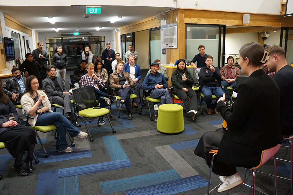 A gathering of 20-30 people in a room listening to two speakers sitting on stools
