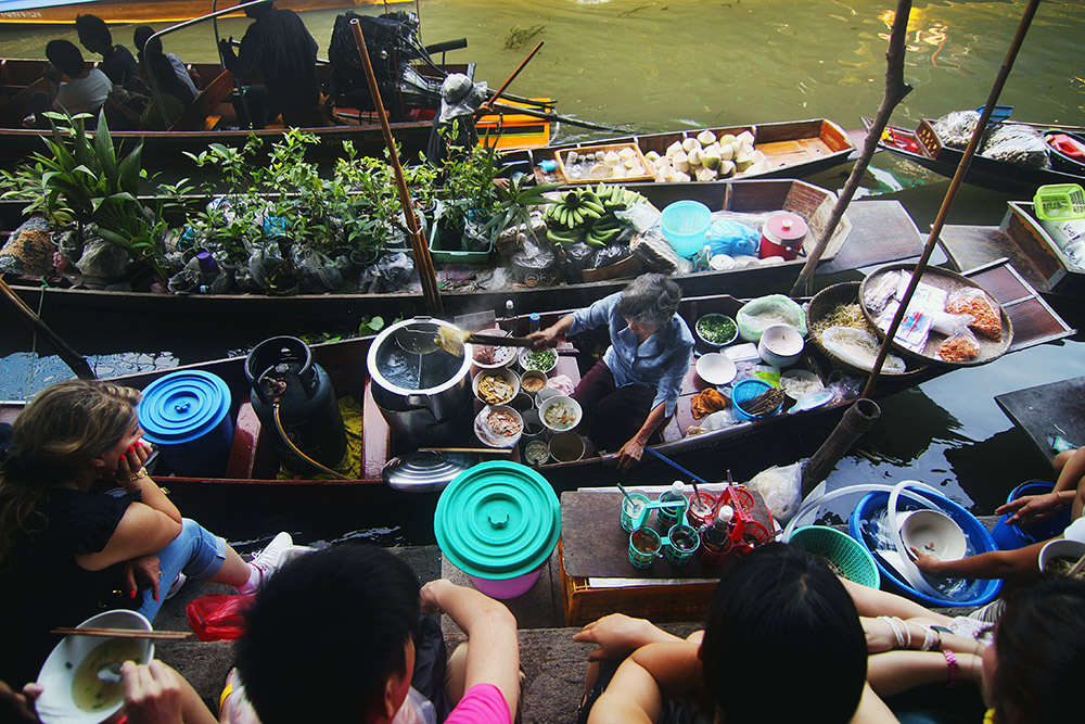 A woman serving soup from a boat to people sitting on a canal side