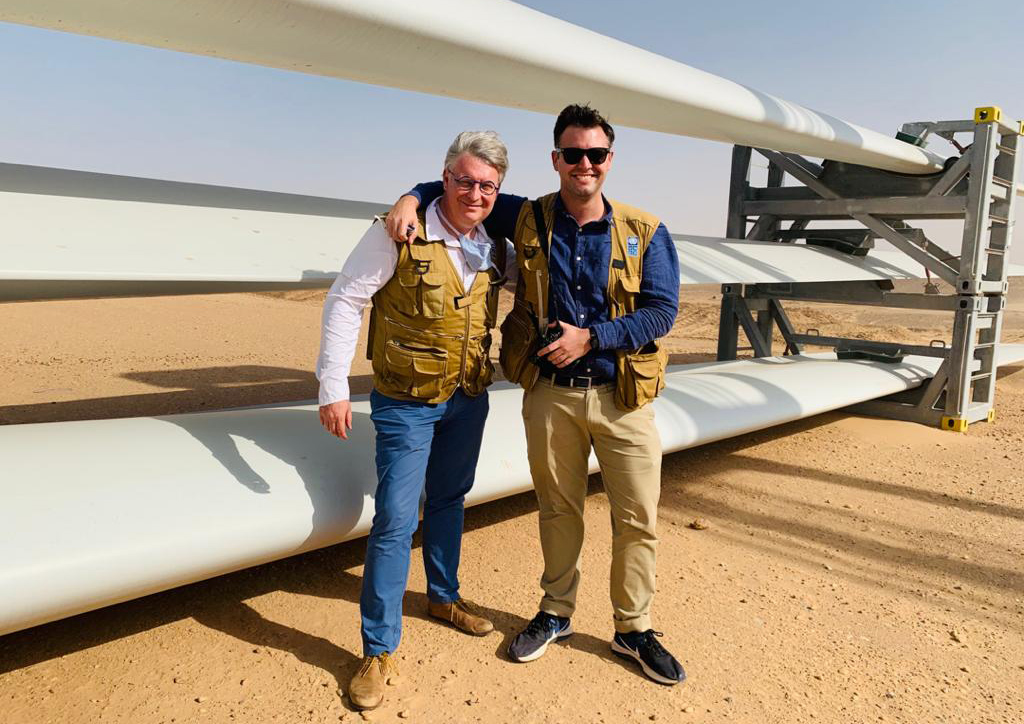 Will in the desert with his arm around a man and the bladesof a wind turbine behind him