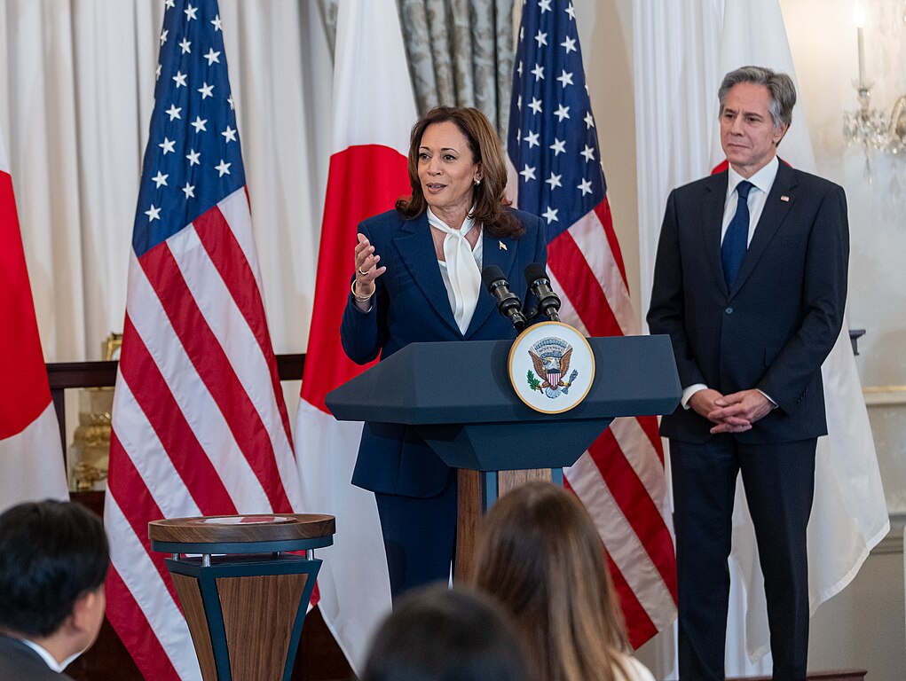 Vice President Kamala Harris adressing a room from a lecturn with the American and Japanese flags behind her and watched on by Secretary of State Anthony Blinken