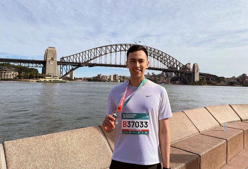 Wesley standing in front of the Sydney Harbour Bridge holding a medalion after completing a running race
