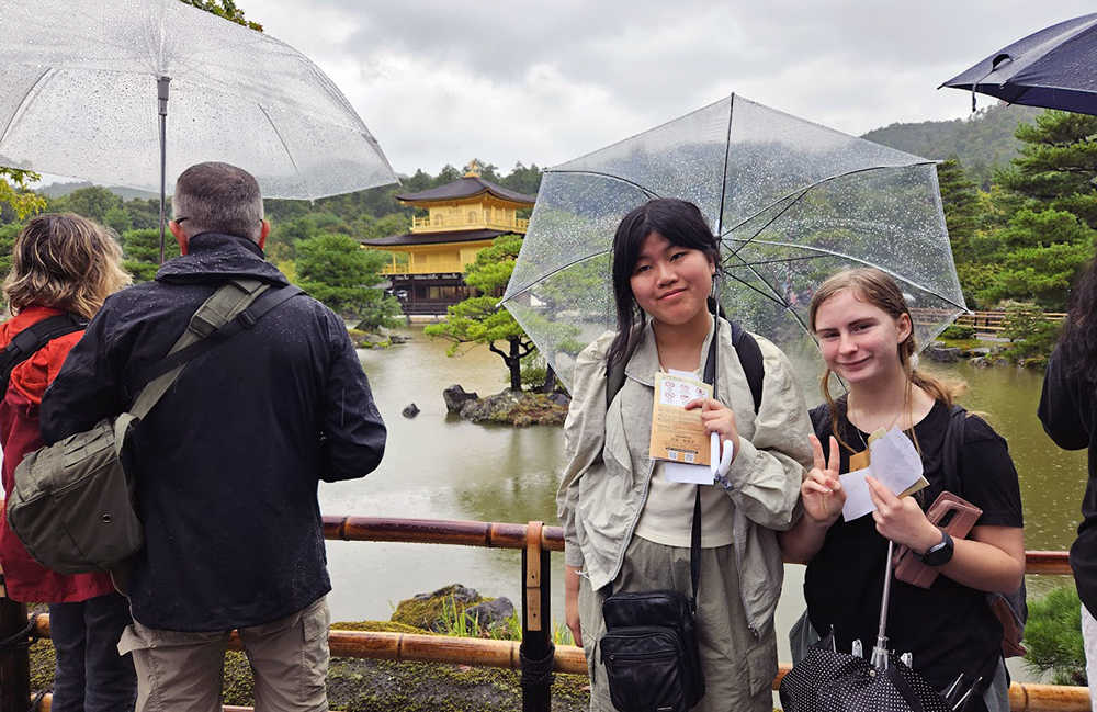Two students standing under an umbrella with Shinkakuji, the Golden Temple, behind them