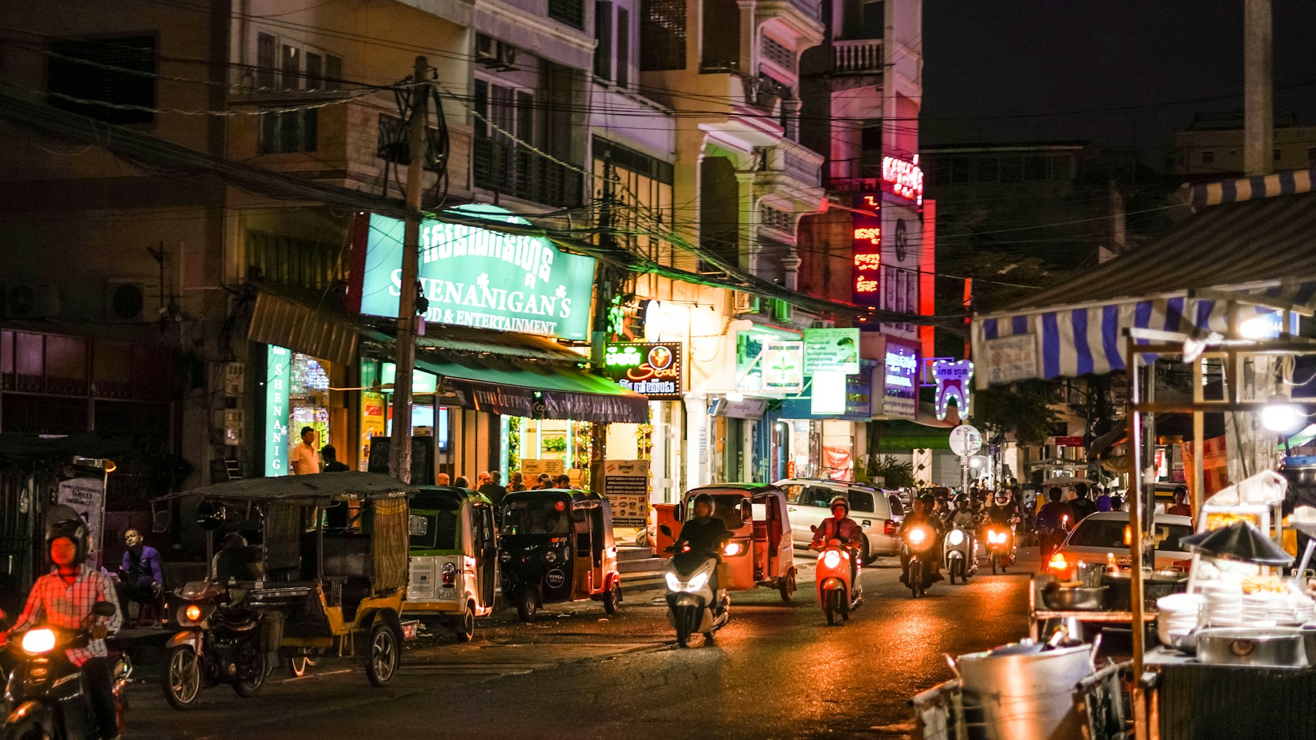 a street at night with neon sings, scooters and cars