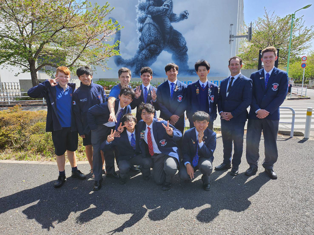 A group of 11 students and a teacher posing in front of a Godzilla mural