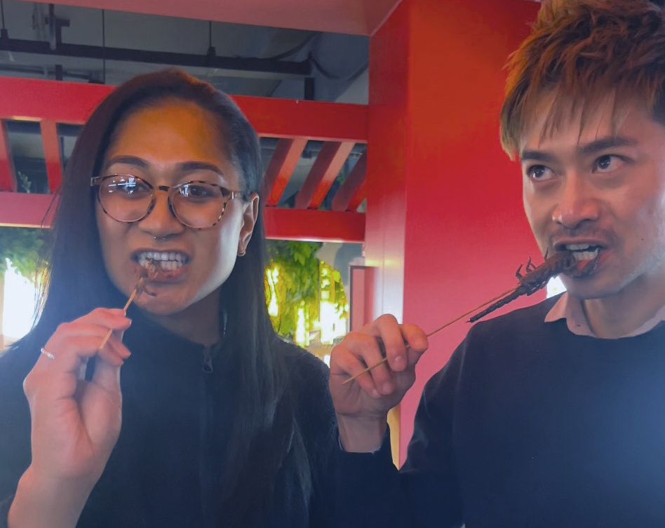 Two Leadership Network members, Shalini and Max, biting into deep-fried scorpions