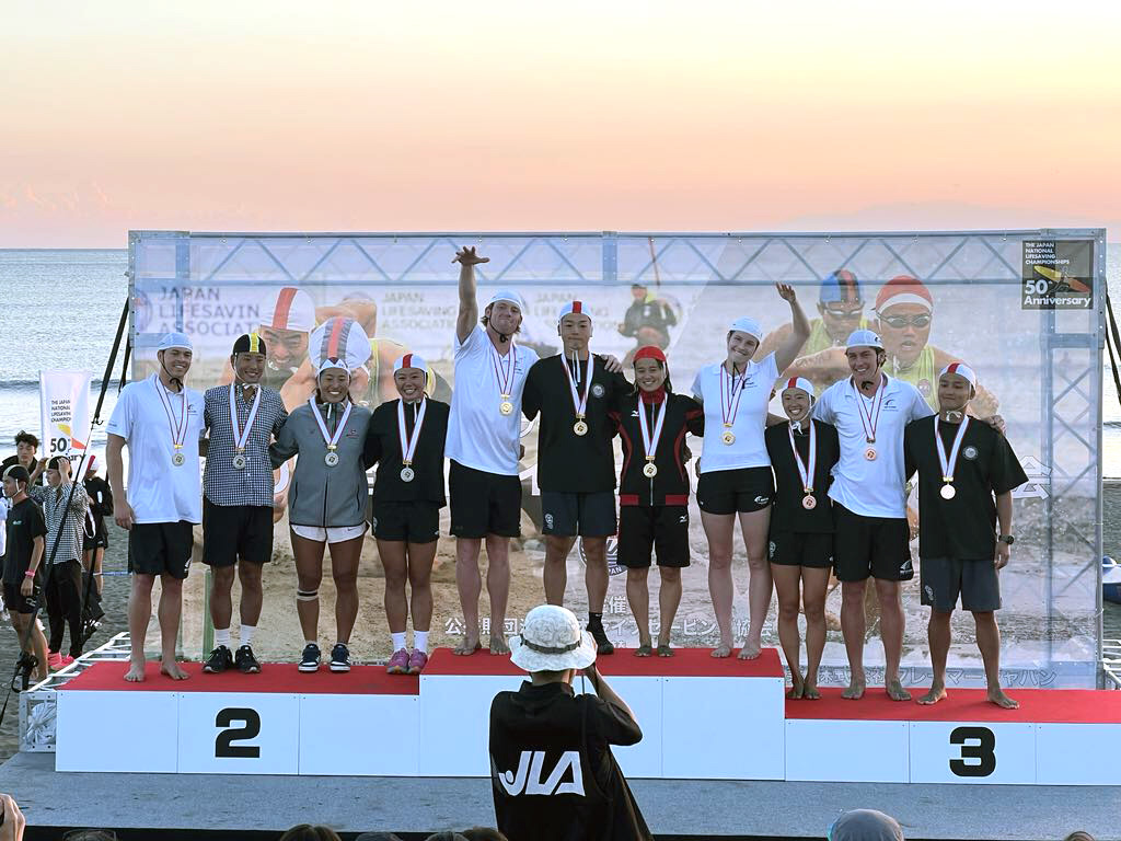 Eleven compettors standing on a podium on a beach at sunset