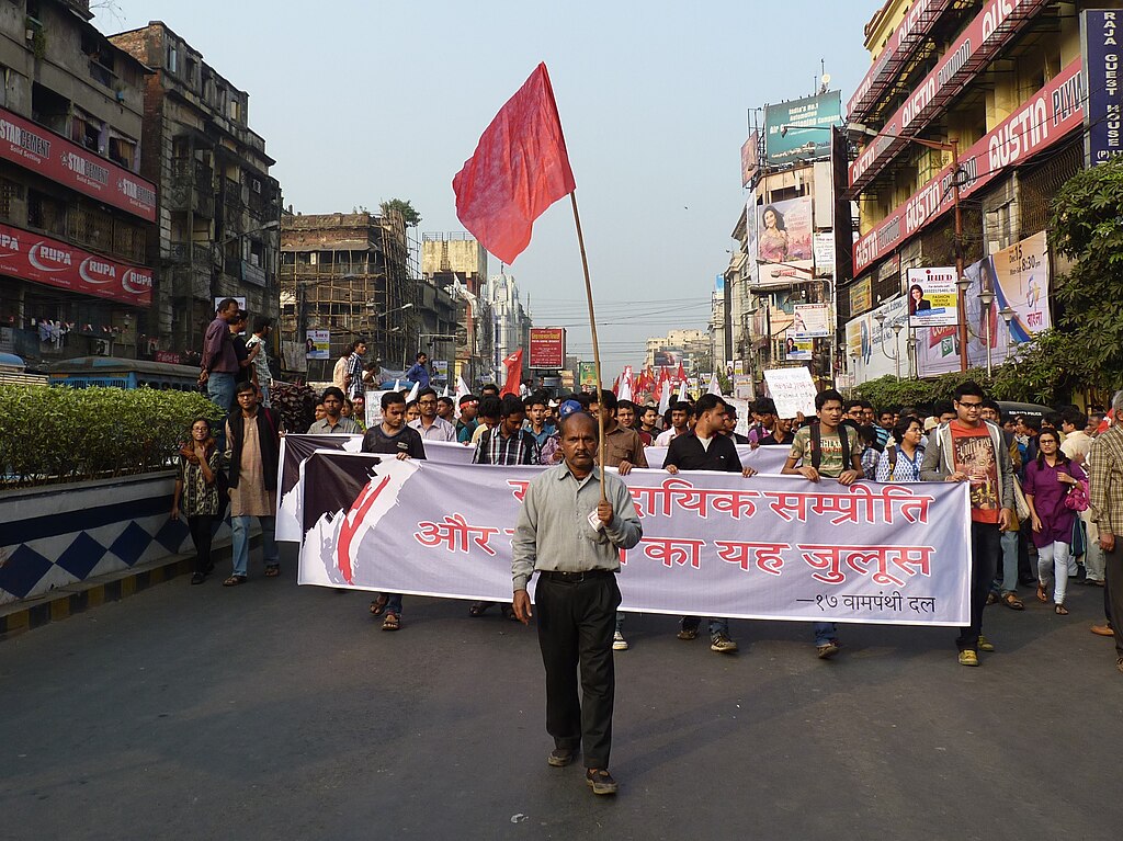A man carrying a red flag in front of a procession carrying banners in an Indian street