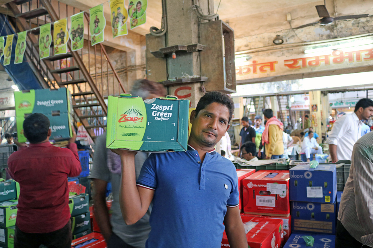 An Indian man in a market holding a box of kiwifruit on his shoulder