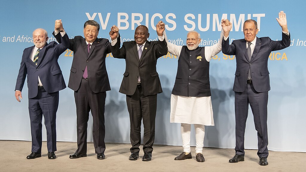 Five leaders of BRICS countries standing hand-in-hand in front of a BRICS banner