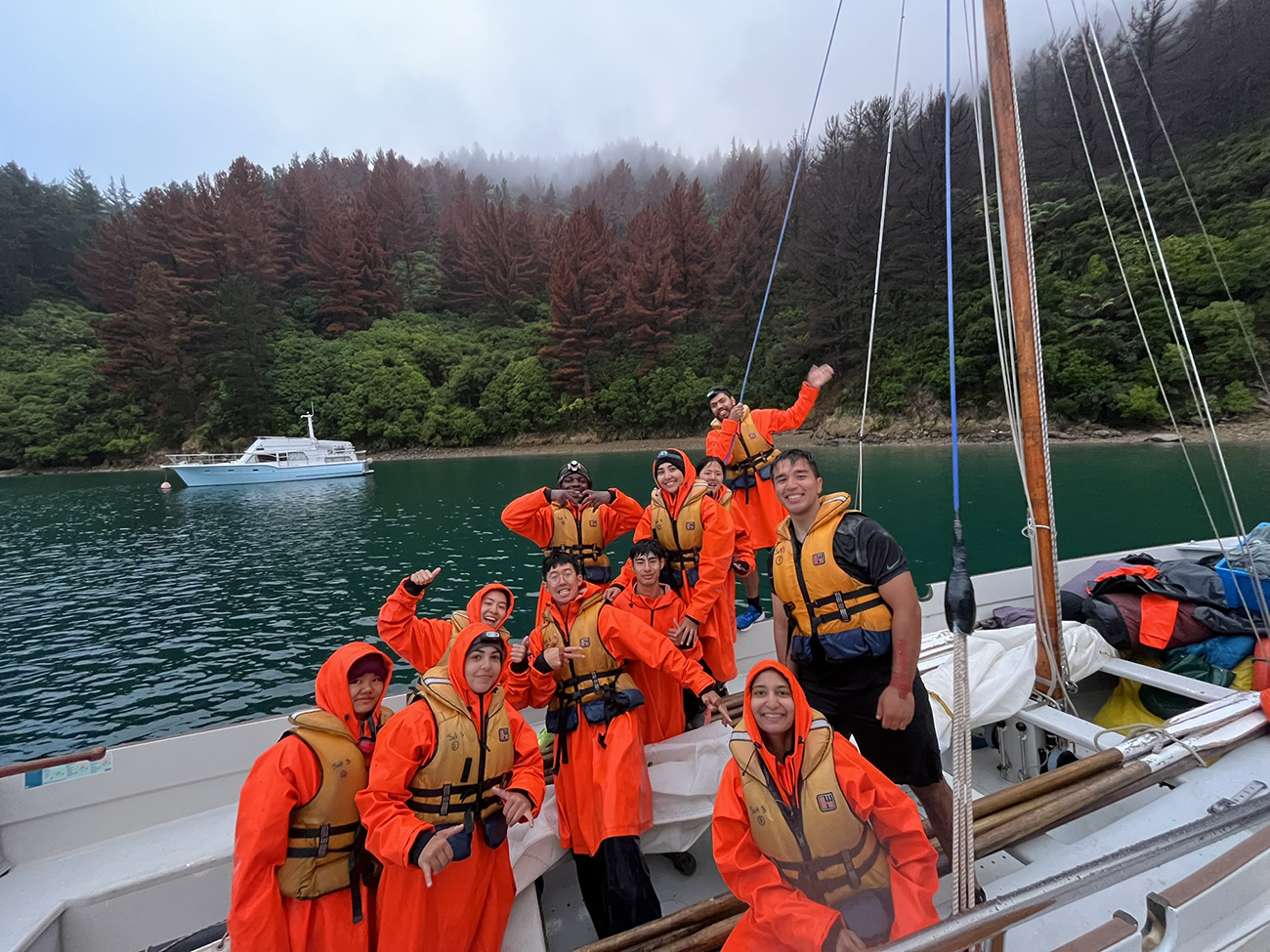 A group of Outwardbound participants dressed in bright orange wet weather gear on a yacht with misty pine-clad hills behind them
