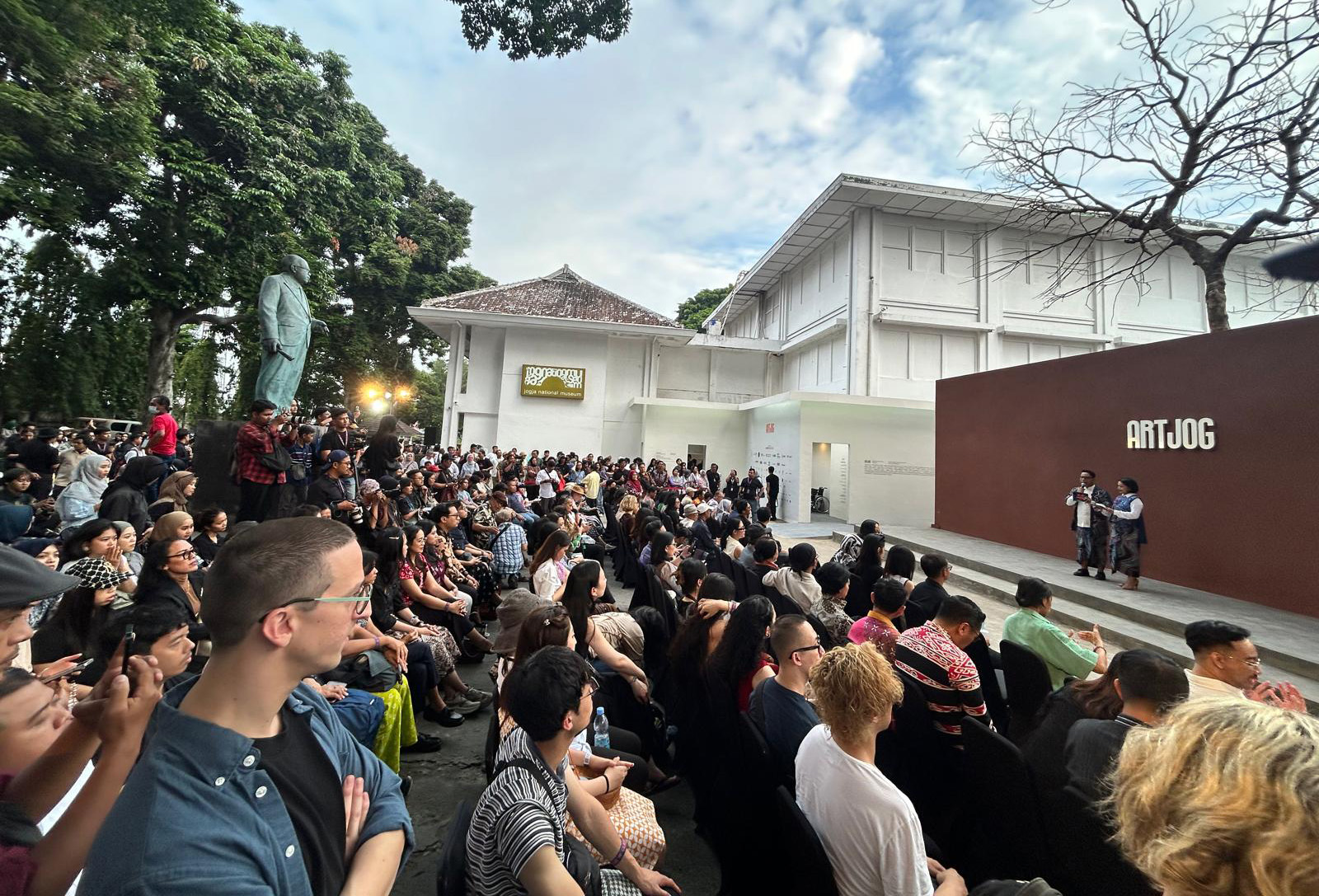 A audience sits outside on chairs as two people present on steps outside the ARTJOG exhibition space