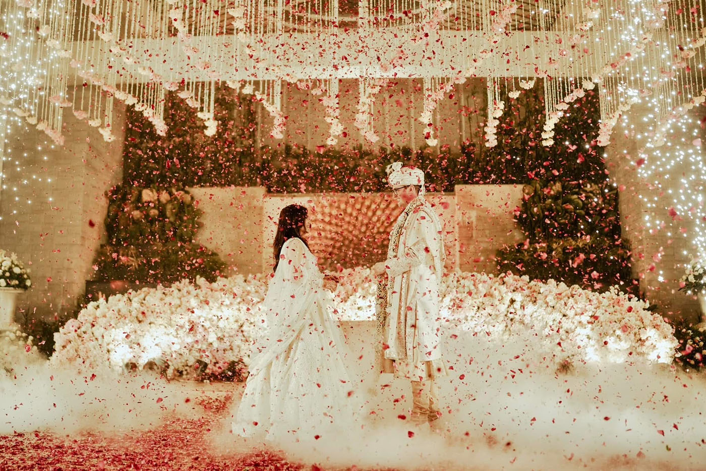 Brent and Anisha standing on stage in their white wedding attire with petals falling around them