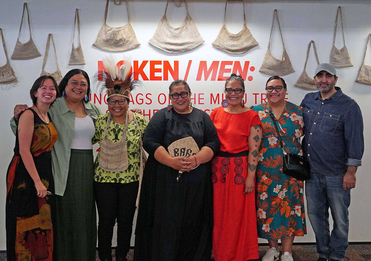 Seven people lined up for a group photo in front of a wall of noken - bags made of bark in Papua New Guinea