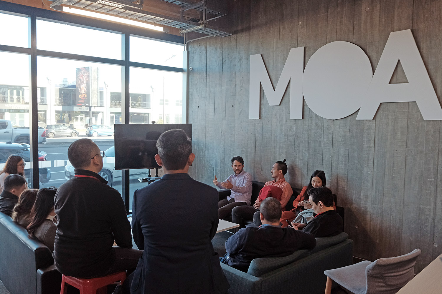 The entrepreneurs listening to a man speak in an office with a large sign  reading MOA on the wall behind him