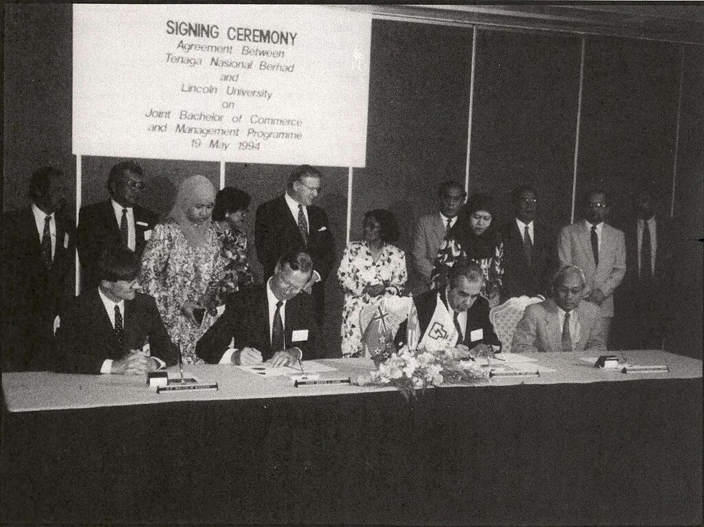 People sitting at a table signing documents with others standing behind them, including former New Zealand Prime Minister Jim Bolger