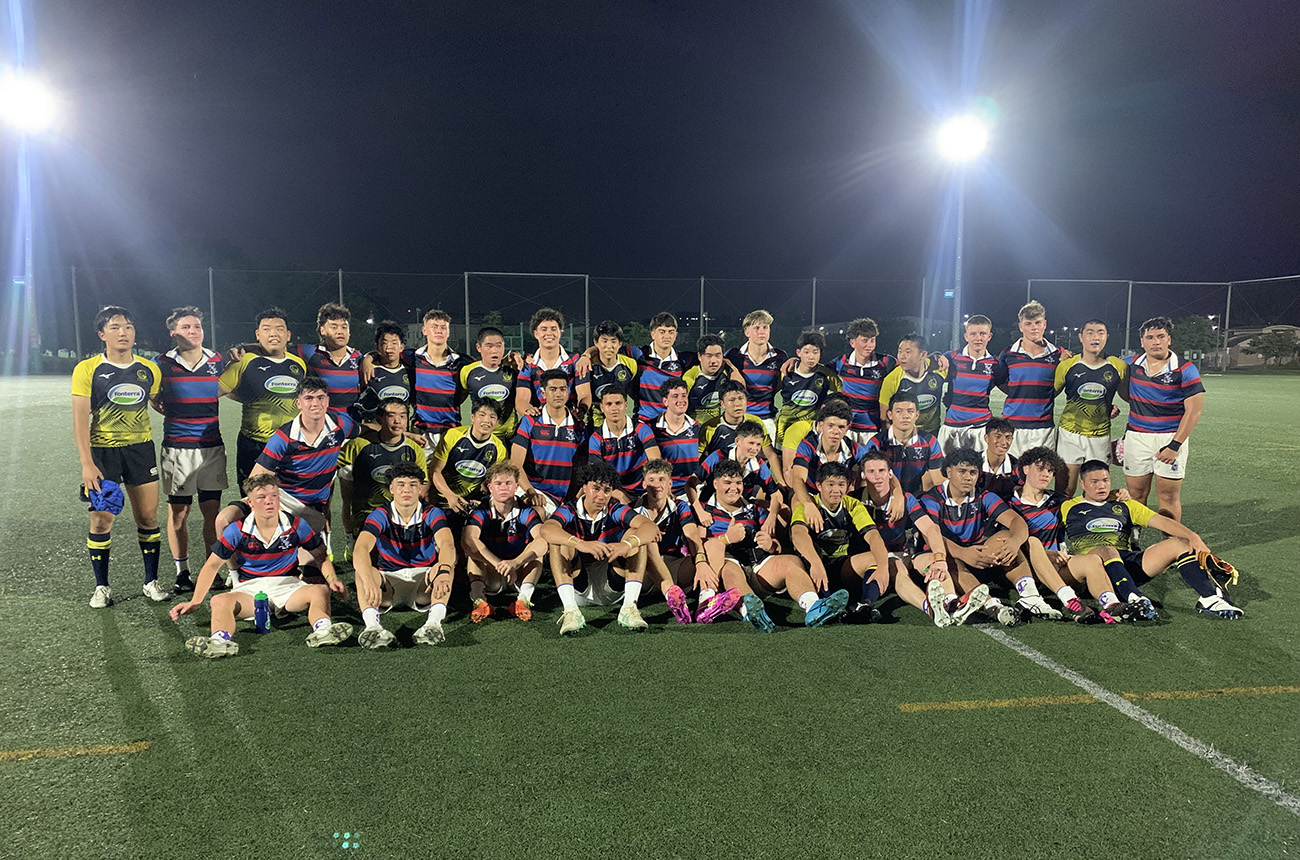 The two rugby teams pose for a group photo on a field, lit by the ground's spotlights