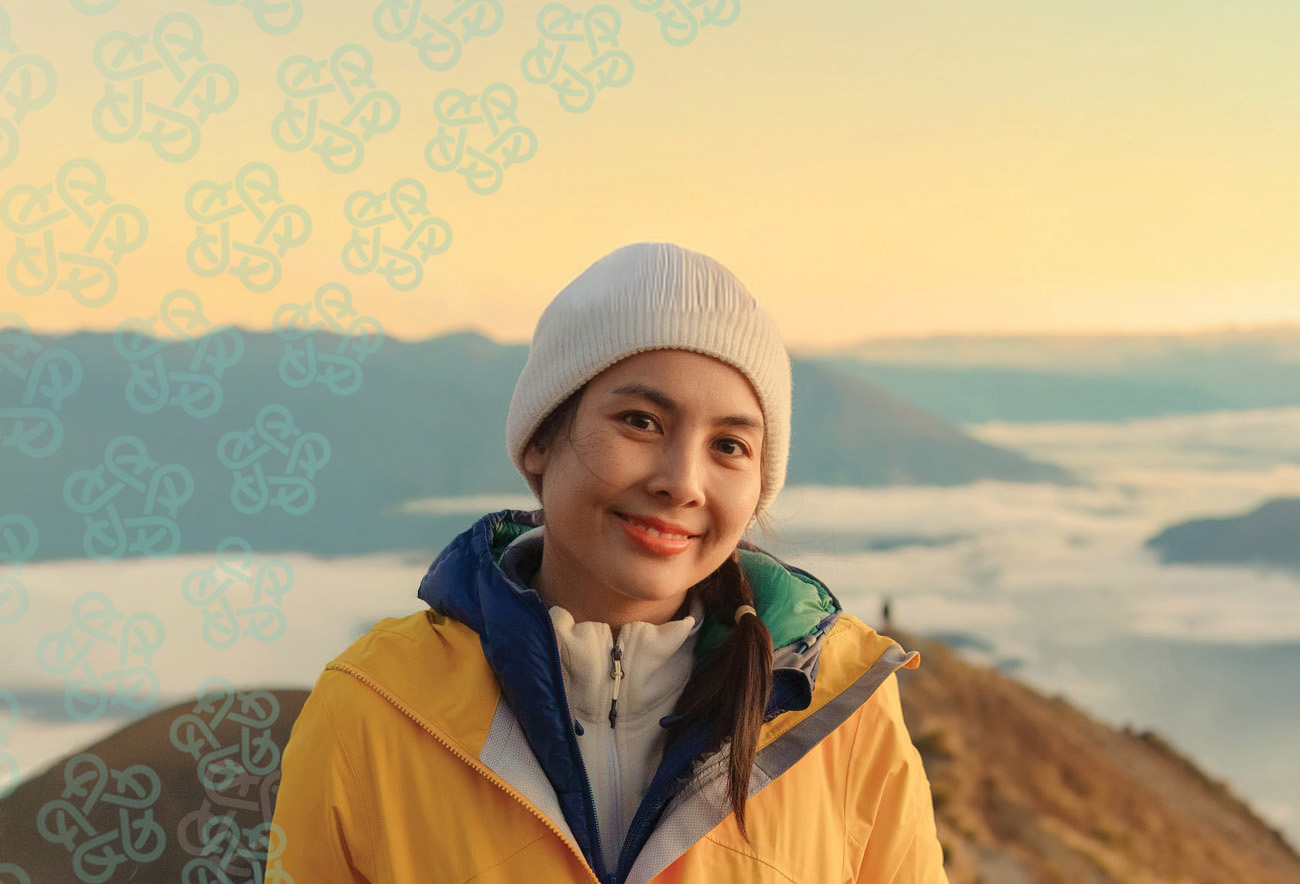 A woman wearing a beanie and jacket posing for a photo on a hilltop with cloud-covered hills behind her