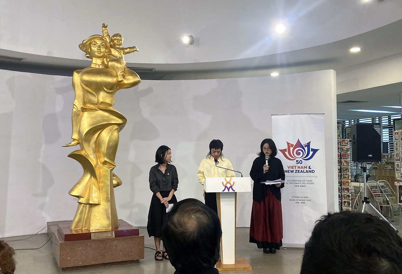 Three people speaking to an audience in a gallery with a large golden scupltute beside them