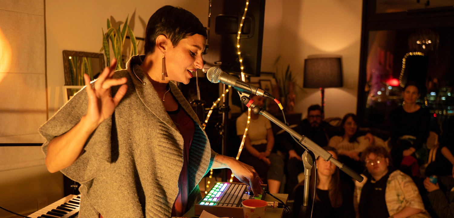 Nadia speaking into a microphone while adjusting dials on a mixing board, while being watched by a small gathering of people on a