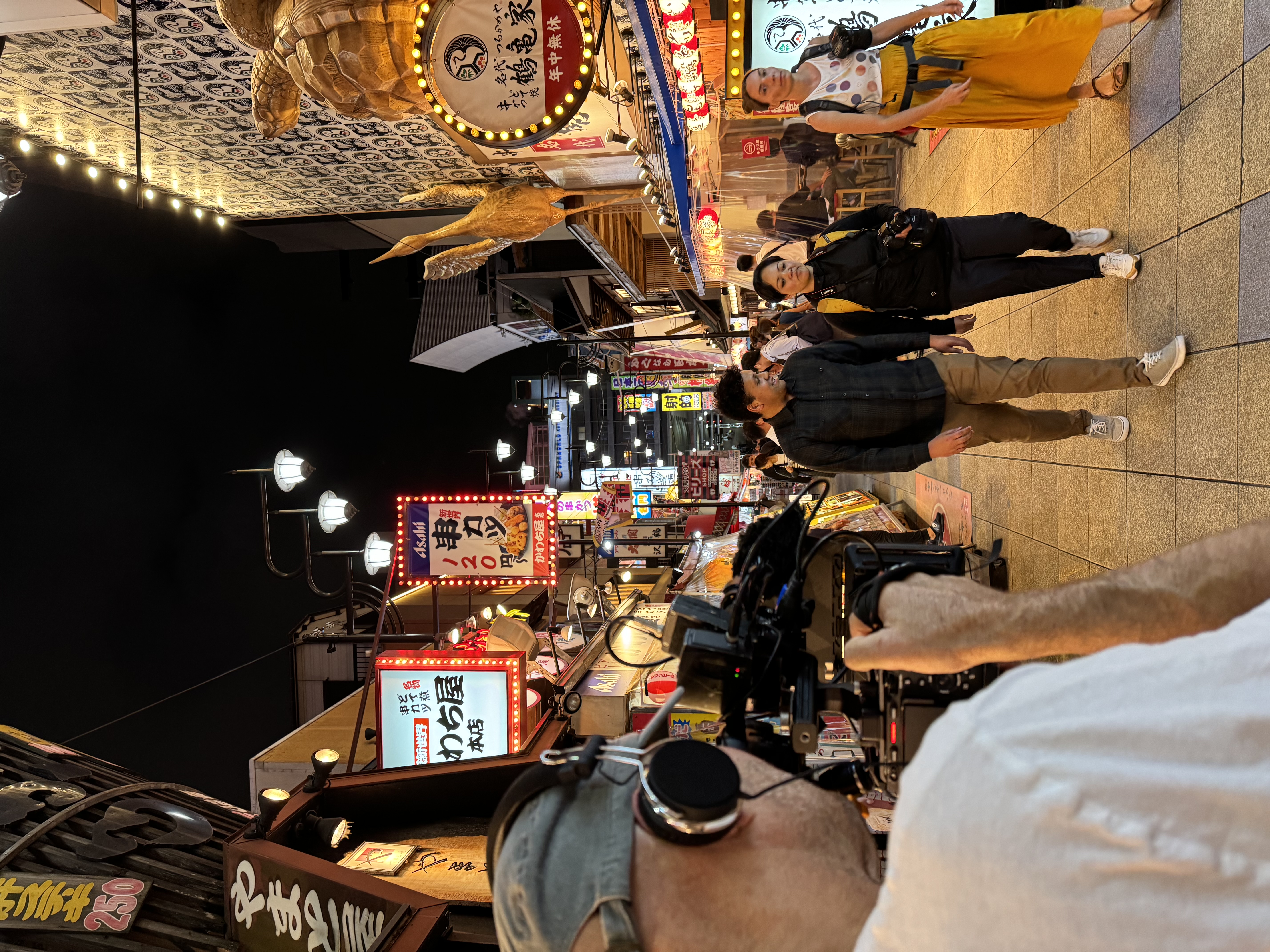 A cameraman filming three people walking through a busy walking street in Japan at night, lit up by shop signs and lanterns