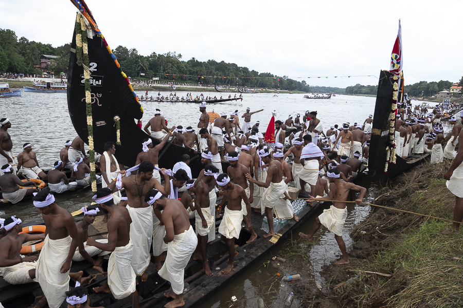 A crowd of men dressed in white cloths clamber over boats on a river.