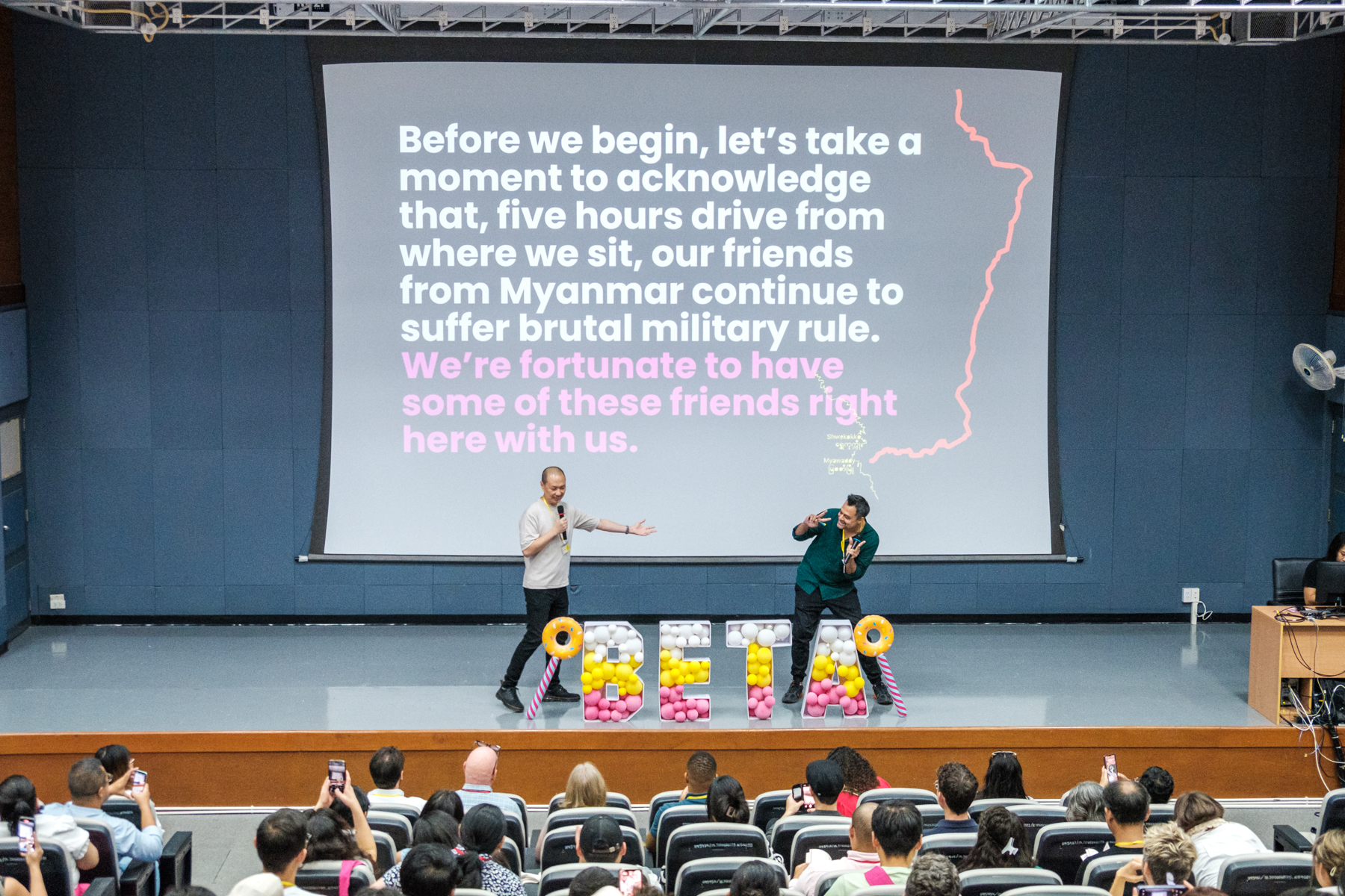 Two men on a lecture hall stage hold microphones and gesture towards each other, with a large Powerpoint presentation in the background