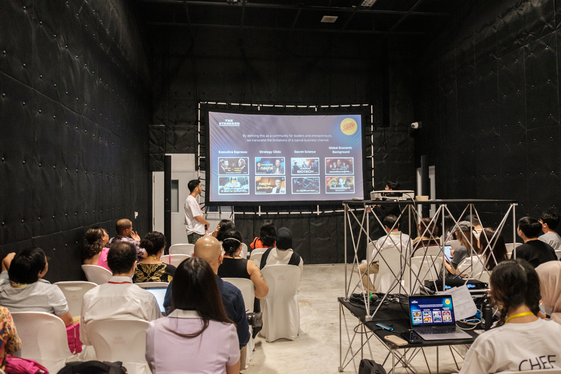 An audience sits in plastic chairs, looking at a presentation set against a large black backdrop