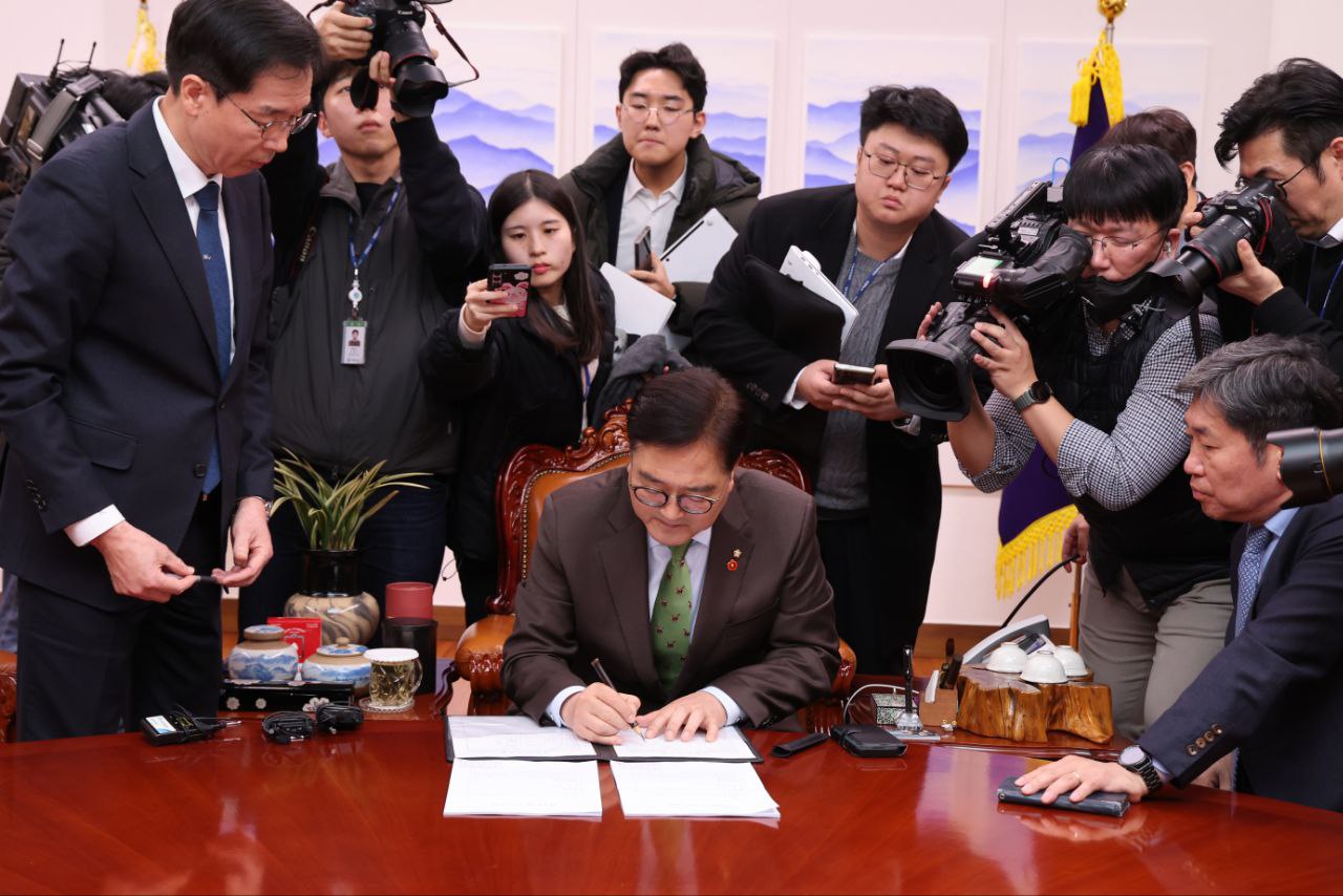 A man sitting at a desk leans forward to sign paperwork. He is surrounded by journalists holding cameras, recording devices and notepads.