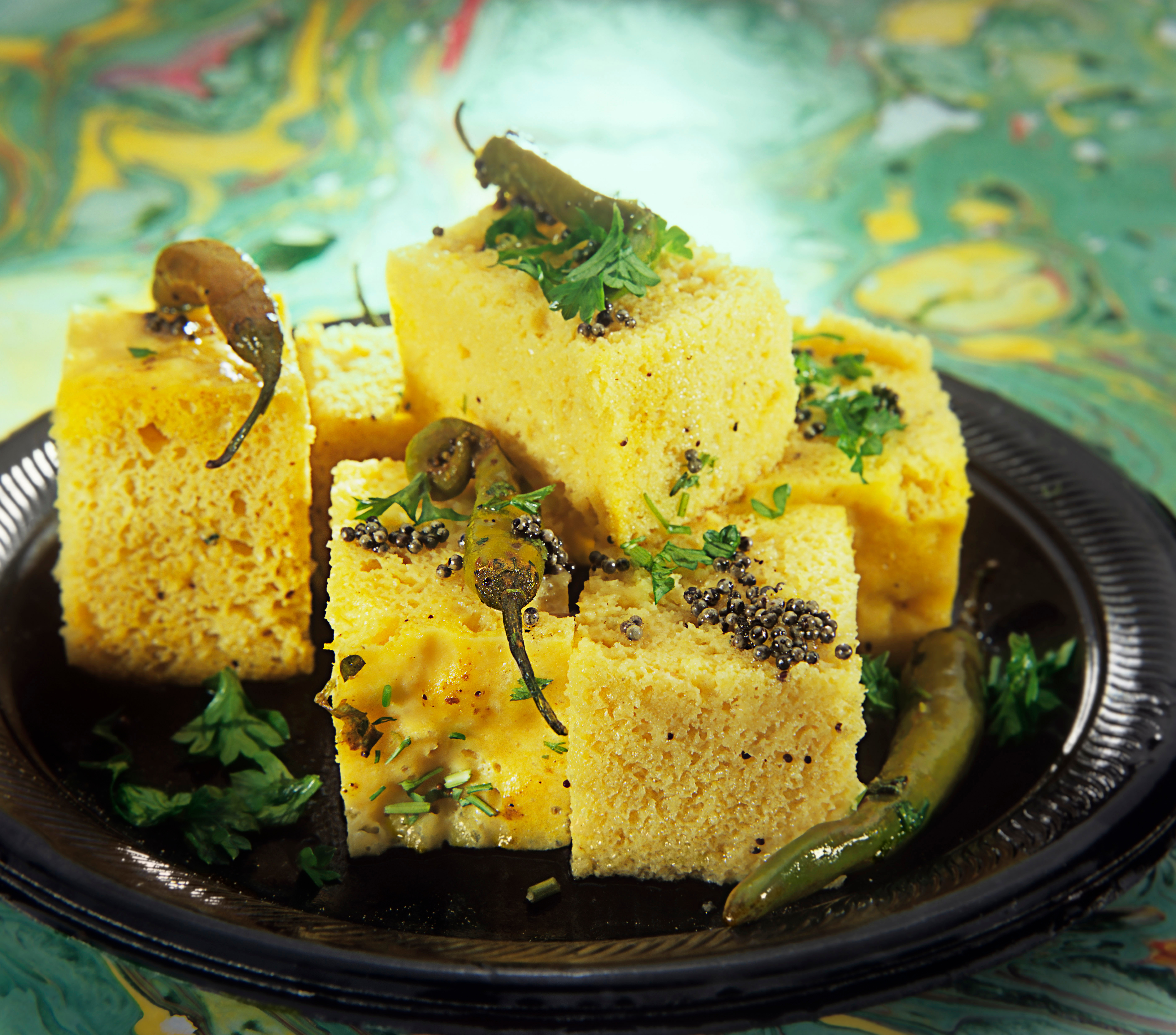 Fluffy cubes of a sponge-like food sit on a plate, topped with green chillies.