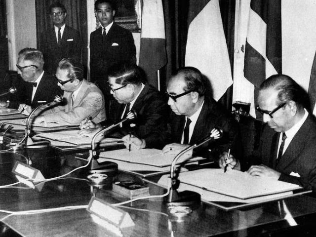 A black and white photo of five suited men sitting behind a desk, all signing papers.